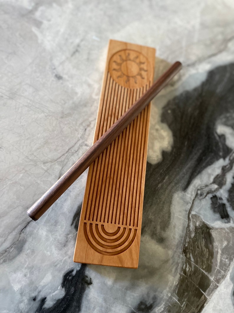 May include: A wooden rolling pin with a sun and rainbow design. The rolling pin is made of light wood and has a dark wood handle. The sun and rainbow design is carved into the wood.