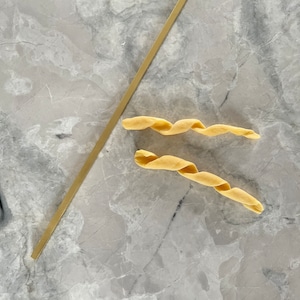 May include: A top-down view of uncooked pasta and a golden-colored metal tool on a gray marble surface. Two pieces of twisted pasta are next to a long, thin, rectangular tool. The pasta is a light yellow color.