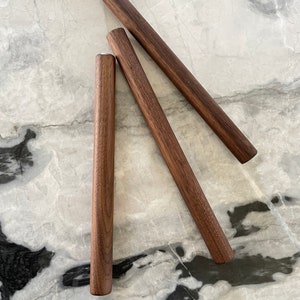 May include: Three wooden dowels, each about 12 inches long, are arranged in a triangular shape on a white marble surface.