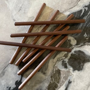 Puede incluir: Un conjunto de siete clavijas de madera, dispuestas en forma de cruz sobre una superficie de mármol. Las clavijas son de color marrón oscuro y tienen un acabado liso y pulido.