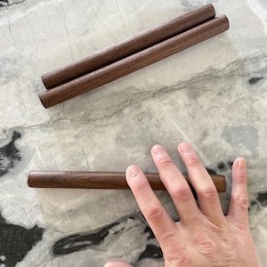 May include: Two brown wooden dowels, one on top of the other, and a single brown wooden dowel lying on a white marble surface.
