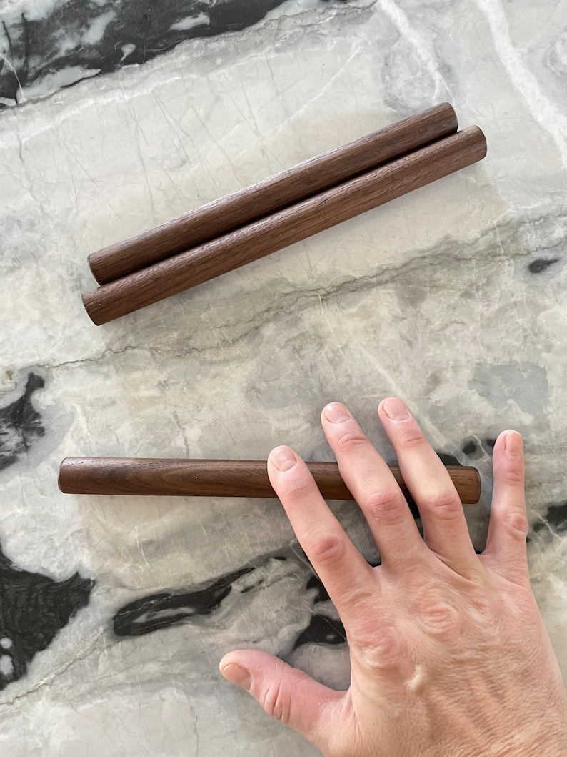 May include: Two brown wooden dowels, one on top of the other, and a single brown wooden dowel lying on a white marble surface.