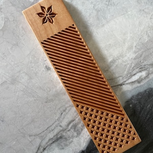 Pode incluir: Um marcador de p&aacute;gina de madeira com um design de flor esculpido na parte superior e um padr&atilde;o geom&eacute;trico na parte inferior. O marcador de p&aacute;gina &eacute; de cor castanho claro e tem um acabamento liso.