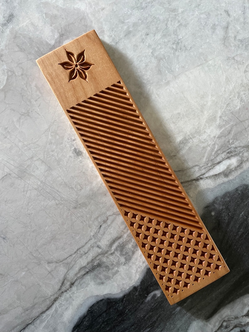 Pode incluir: Um marcador de p&aacute;gina de madeira com um design de flor esculpido na parte superior e um padr&atilde;o geom&eacute;trico na parte inferior. O marcador de p&aacute;gina &eacute; de cor castanho claro e tem um acabamento liso.