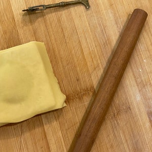 May include: A wooden rolling pin and a small, brass tool on a wooden cutting board. The rolling pin is brown and the tool is gold.