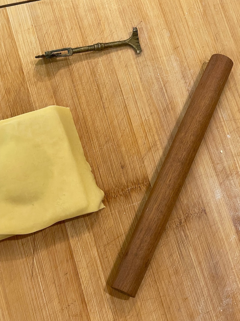 May include: A wooden rolling pin and a small, brass tool on a wooden cutting board. The rolling pin is brown and the tool is gold.