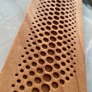 May include: A close-up of a wooden board with a pattern of small, round holes cut into the surface. The holes are arranged in a curved line, creating a wave-like effect. The wood is a light brown color.