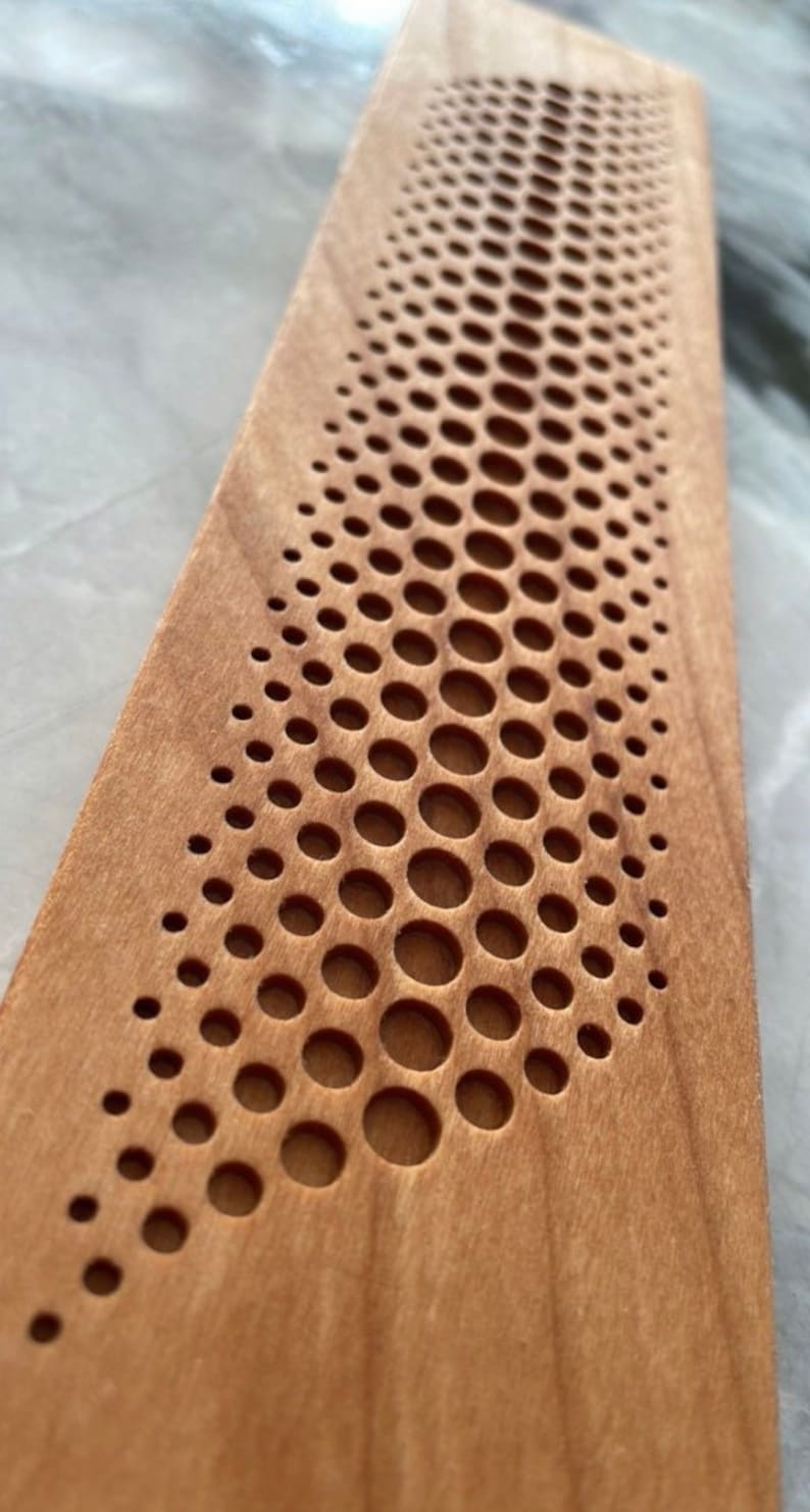 May include: A close-up of a wooden board with a pattern of small, round holes cut into the surface. The holes are arranged in a curved line, creating a wave-like effect. The wood is a light brown color.
