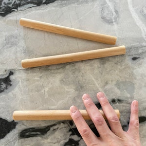 May include: Three wooden dowels, two lying flat and one held by a hand. The dowels are light brown and smooth.