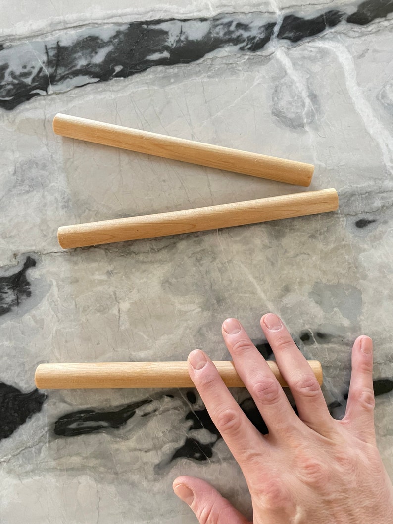 May include: Three wooden dowels, two lying flat and one held by a hand. The dowels are light brown and smooth.