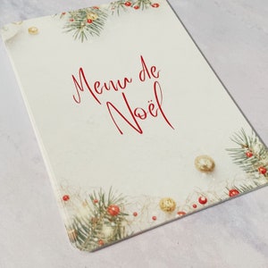 May include: A white card with the words 'Menu de Noël' written in red cursive. The card is decorated with green pine branches, red berries, and gold ornaments.
