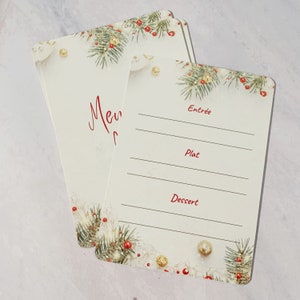 May include: Two blank white cards with a Christmas theme. The cards have a border of green pine branches with red berries and gold ornaments. The top card has the word 'Menu' written in red cursive. The bottom card has the words 'Entree', 'Plat', and 'Dessert' written in red. The cards are perfect for writing down a holiday menu.