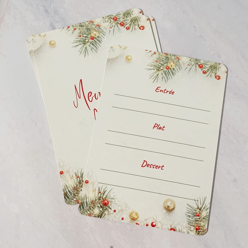 May include: Two blank white cards with a Christmas theme. The cards have a border of green pine branches with red berries and gold ornaments. The top card has the word 'Menu' written in red cursive. The bottom card has the words 'Entree', 'Plat', and 'Dessert' written in red. The cards are perfect for writing down a holiday menu.