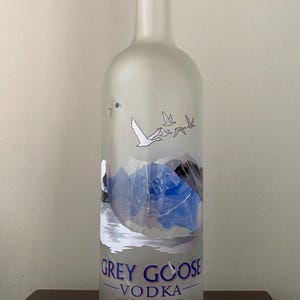 May include: A frosted glass Grey Goose Vodka bottle with a cork stopper. The bottle features a mountain and lake design with flying geese. The text "GREY GOOSE VODKA" is displayed prominently, along with "FRANCE".