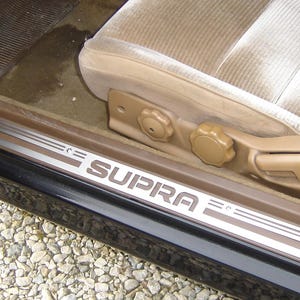 Puede incluir: Interior de coche negro con asientos de cuero marrón y una inscripción "SUPRA" en el umbral de la puerta.