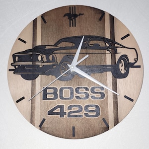 May include: Wooden wall clock featuring a black silhouette of a classic car, with the words "BOSS 429" in bold letters. The clock has a light brown wooden background with vertical stripes and black clock hands. A horse logo is at the top.