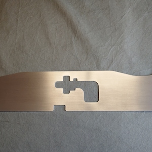 Puede incluir: Un componente de metal cepillado con forma rectangular y varias secciones recortadas. La pieza tiene un color metálico claro y parece estar hecha de una sola lámina de metal. Presenta bordes redondeados y una serie de agujeros y ranuras.