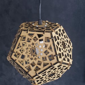 Puede incluir: Lámpara colgante geométrica de madera con intrincados patrones recortados. La luz está suspendida de un cable negro sobre un fondo oscuro. El diseño del accesorio presenta formas geométricas repetidas, creando un elemento de iluminación decorativo.