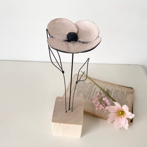 Może przedstawiać: Ręcznie wykonana metalowa rzeźba kwiatu z jasno różowymi płatkami i czarnym środkiem. Kwiat jest zamontowany na drewnianej podstawie z klocków. Stary książka i różowy kwiat są w tle.