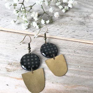 Puede incluir: Un par de pendientes colgantes. Cada pendiente presenta una parte superior circular gris oscuro con un patrón de estrellas, conectada a una parte inferior semicircular de color dorado. Los pendientes cuelgan de ganchos para las orejas de color bronce. Flores blancas en el fondo.