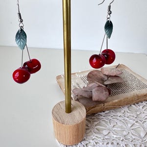 Peut inclure: Boucles d'oreilles cerises avec des cerises rouges brillantes et des feuilles vertes. Elles sont présentées sur un support doré avec une base en bois. Le fond comprend un livre vintage et de la dentelle blanche.