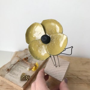 Op de afbeelding: Een handgemaakte, geelgouden bloemensculptuur met een zwart centrum, gemonteerd op een houten blok. De bloem heeft een metallic glans en wordt gepresenteerd op een houten oppervlak. De sculptuur is een uniek decoratief item.