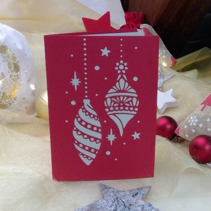 Puede incluir: Una tarjeta de Navidad roja con un diseño recortado blanco de dos adornos colgantes. Los adornos están decorados con espirales y puntos blancos. La tarjeta está rodeada de artículos decorativos blancos, dorados y marrones.