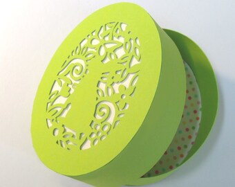 May include: A green round box with a cut-out design of flowers and swirls. The box is open and reveals a white round disc with colorful polka dots.