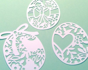 May include: Three white paper cut-out ornaments in the shape of Easter eggs. The ornaments are decorated with floral designs, a bow, and a heart.
