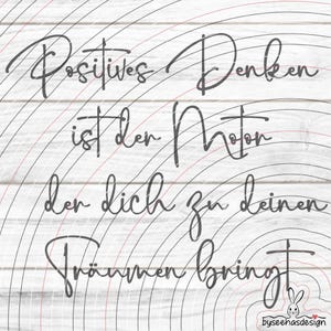 以下が含まれることがあります： 白い木製の背景に、暗い灰色のスクリプトフォントで「Positives Denken ist der Motor der dich zu deinen Träumen bringt」というテキスト。 デザインには、黒と赤の同心円が含まれています。