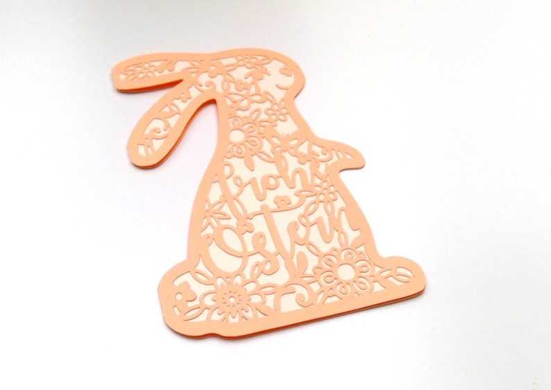 May include: A peach-colored paper cutout of a bunny rabbit with intricate floral designs and the text "Frohe Ostern" inside the cutout.