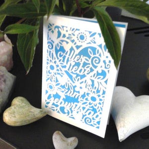 May include: A white greeting card with a blue background and intricate floral cutouts. The card features the German text "Alles Liebe zum Geburtstag". Decorative heart-shaped stones and greenery surround the card.