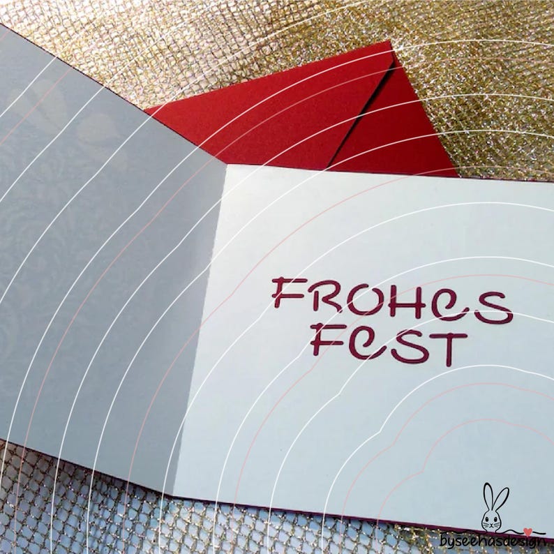 May include: A folded greeting card with a white interior featuring the dark red text "FROHES FEST" in a stylized font. The card is positioned against a red envelope, with a small black rabbit logo and the text "byseehasdesign" at the bottom.