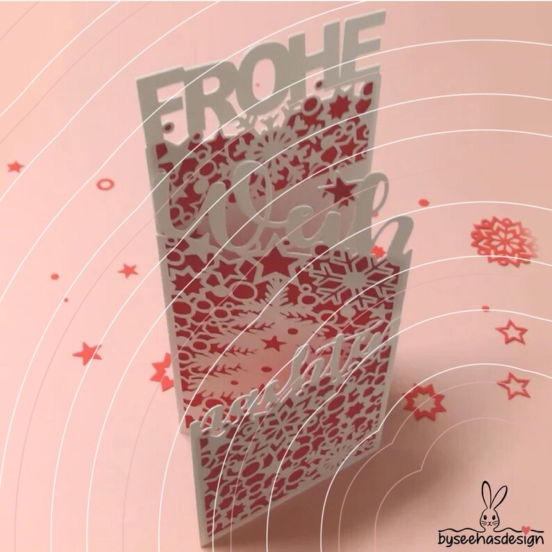 May include: A vertical greeting card featuring intricate laser-cut patterns of snowflakes and stars in white and red. The text FROHE WEIHNACHTEN is cut out from the top and middle sections. Small red star and snowflake confetti surround the card.