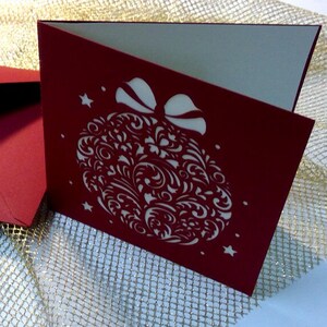 May include: A deep red folded greeting card featuring a white laser-cut design of a Christmas ornament with intricate swirling patterns and a bow at the top, accented by small star shapes on the front cover, accompanied by a matching red envelope.