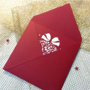May include: A deep red envelope featuring a triangular flap with a central die-cut design. The cutout reveals a white interior layer decorated with an intricate, repeating floral pattern and a bow shape at the top, creating a layered effect.