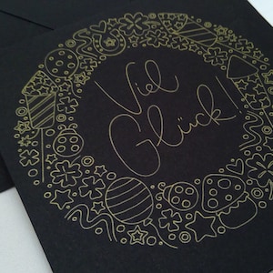 Op de afbeelding: Een zwarte kaart met een gouden omtrek van een cirkel gevuld met feestelijke krabbels. De tekst "Viel Glück!" is in gouden cursiefschrift in de cirkel geschreven.