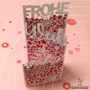 May include: A vertical laser-cut greeting card featuring a red and white layered design with intricate snowflake and star patterns. The card displays the German text FROHE WEIHNACHTEN in bold, die-cut lettering integrated into the structure.