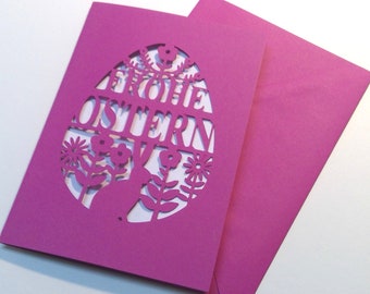 May include: A pink card with a cut-out design of an Easter egg with the words "Frohe Ostern" inside. The egg is surrounded by flowers and a bunny rabbit.