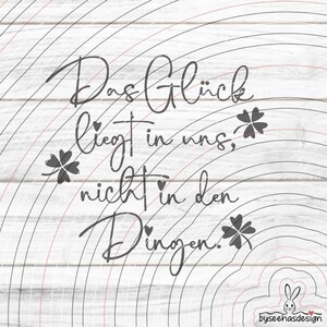May include: A digital design with the German text "Das Glück liegt in uns, nicht in den Dingen" in a script font, meaning "Happiness lies within us, not in things." The design includes four-leaf clovers and a rainbow-like background.