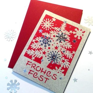May include: A festive Christmas card with a red background and a white snowflake design. The card features the German text "FROHES FEST" in red. A red envelope is behind the card, with silver star and snowflake accents.