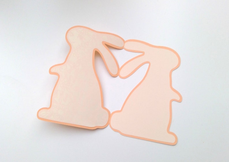 May include: Two peach-colored paper bunny cutouts facing each other, forming a heart shape.