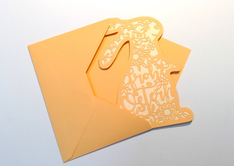 May include: A peach-colored card with a cutout of a bunny rabbit. The bunny is decorated with floral designs and the text "Frohe Ostern" is written inside the bunny.
