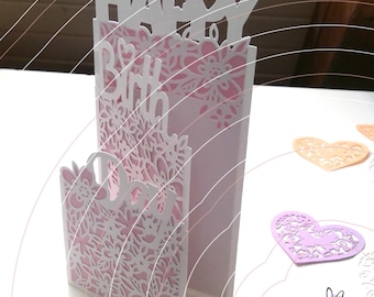 May include: A white laser-cut paper greeting card with a layered floral design and the text HAPPY BIRTH DAY. The card features intricate cutouts of flowers and hearts, revealing a soft pink background layer within the structure.
