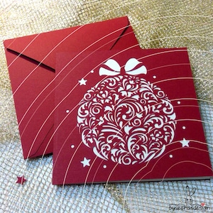 May include: A square red greeting card featuring a white die-cut Christmas ornament design with intricate floral patterns and a bow at the top, accompanied by a matching red envelope. Small white stars are scattered around the ornament.