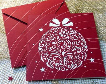 May include: A square red greeting card featuring a white die-cut Christmas ornament design with intricate floral patterns and a bow at the top, accompanied by a matching red envelope. Small white stars are scattered around the ornament.