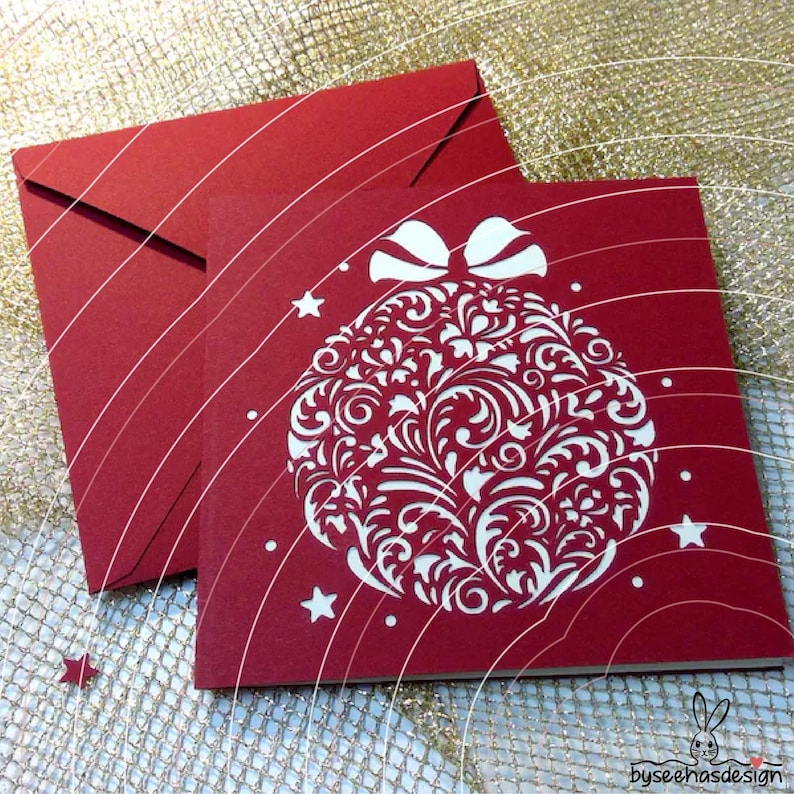 May include: A square red greeting card featuring a white die-cut Christmas ornament design with intricate floral patterns and a bow at the top, accompanied by a matching red envelope. Small white stars are scattered around the ornament.