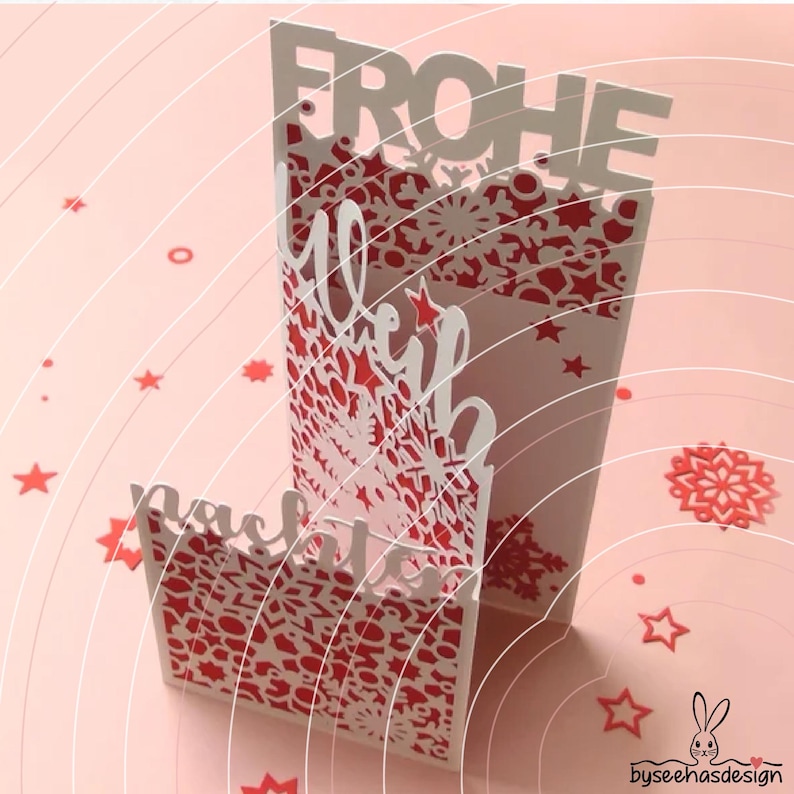 May include: A white and red laser-cut paper card featuring the German text FROHE WEIHNACHTEN. The design includes intricate snowflake and star patterns cut into the paper, with a layered structure that creates depth and a festive appearance.