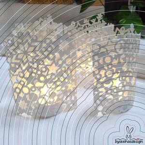 May include: Two cylindrical white paper luminaries featuring intricate laser-cut patterns of snowflakes, stars, and circular shapes. The cutouts allow warm light to glow from within, creating a decorative display with a delicate, lace-like appearance.