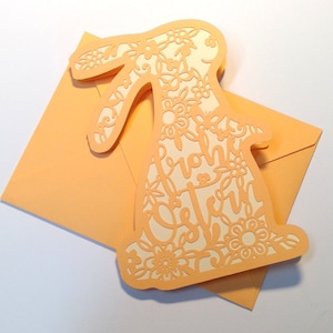 May include: A peach-colored card with a white bunny cutout. The bunny is decorated with floral designs and the text "Frohe Ostern" is inside the cutout.